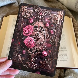 May include: A vintage-style book cover with an ornate gold frame and a red jewel at the top. The frame is adorned with pink roses and vines. The background is dark and mysterious.