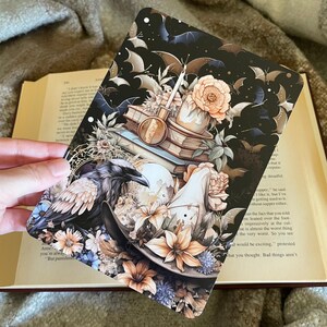 May include: A black raven perched on a stack of books with a candle and flowers. The background is black with bats flying around. The image has a dark and mysterious feel.