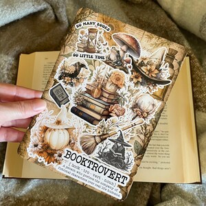 Könnte beinhalten: Eine Sammlung von Aufklebern mit einer Hexe, einem Kürbis, einem Stapel Bücher, einem Frosch, einem Pilz und einem Besen. Die Aufkleber befinden sich auf einem braunen Hintergrund mit dem Text "SO MANY BOOKS" und "SO LITTLE TIME". Der Text "BOOKTROVERT" ist definiert als "Eine Person, die die Gesellschaft von fiktiven Charakteren gegenüber realen Menschen bevorzugt."
