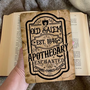 Op de afbeelding: Verouderd bord met de tekst "Old Salem Apothecary, Est. 1845, Enchanted Potions, Tonics, Elixirs." Het bord heeft een zwarte rand en een schedel- en vleermuisafbeelding. Het bord wordt voor een open boek gehouden.