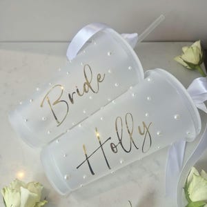 May include: Two white, frosted plastic tumblers with lids and straws. One reads "Bride" in gold script, the other "Holly". Both are decorated with small, pearl-like embellishments and white ribbon, perfect for a celebration.