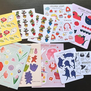 Puede incluir: Una colección de hojas de pegatinas con varios diseños. Las hojas muestran ilustraciones de plantas, fresas, gatos y elementos de Halloween. La paleta de colores incluye pasteles, con toques de rojo, azul y amarillo. Algunas hojas tienen texto como "Happy Strawberries" y "Spooky!"