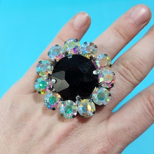May include: A large, black, faceted ring with a cluster of iridescent rhinestones around the center stone.