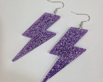 Könnte beinhalten: Ein Paar lila Glitzer-Blitz-Ohrringe. Die Ohrringe haben silberfarbene Haken und bestehen aus einem glitzernden Material. Die Ohrringe haben die Form eines Blitzes.
