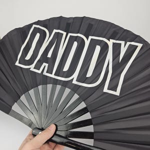 May include: Black hand fan with the word "DADDY" in large white letters. The fan is open, revealing the wooden ribs and handle. The fan is held by a person.