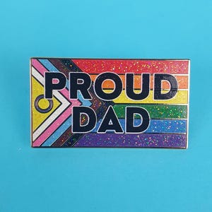 May include: A rainbow glitter enamel pin with the text "PROUD DAD" in black letters. The pin features a rainbow flag design with a gender-inclusive symbol.