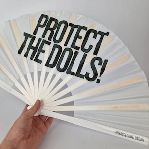 May include: A white folding fan with the text "PROTECT THE DOLLS!" in bold black letters. The fan has wooden ribs and a white handle. The text "@DRAGGEDOUTLONDON" is printed on the handle.