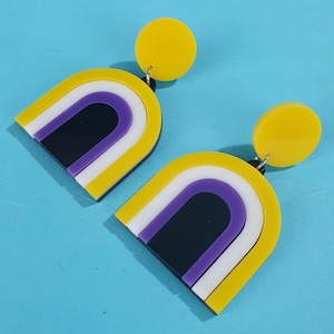 May include: A pair of yellow, white, purple and black acrylic earrings shaped like arches. The earrings have a yellow circle attached to the top.