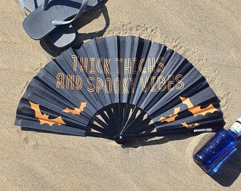 May include: Black folding hand fan with orange text that reads "Thick Thighs and Spooky Vibes". The fan has a bat design and is open on a sandy beach.