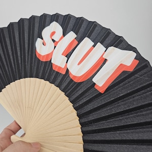 May include: Black folding fan with the word "SLUT" in white letters with a red outline. The fan has a wooden handle and is partially open, displaying the pleated black paper.