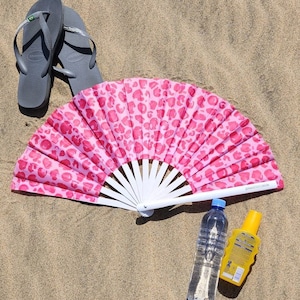 Puede incluir: Un abanico rosa con estampado de leopardo, varillas blancas y mango blanco, abierto sobre arena. Chanclas grises, una botella de agua y un frasco de protector solar también están sobre la arena.