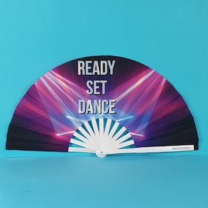 以下が含まれることがあります： 黒と紫のデザインの白い折りたたみ式扇子。扇子には「READY SET DANCE」の文字が印刷されています。