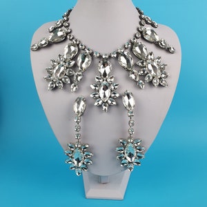 Pode incluir: Um conjunto de colar e brincos de prata com grandes pedras de cristal transparentes. O colar é uma peça chamativa com um design grande e ornamentado. Os brincos são longos e pendentes, com um design semelhante ao do colar.