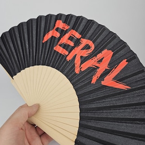 May include: A black folding fan with the word "FERAL" in bright orange lettering. The fan is made of black paper and has a wooden handle. The fan is open and held in a hand.