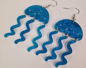 May include: A pair of blue jellyfish earrings. Each earring features a scalloped, semi-circular top with a textured, marbled appearance and wavy, dangling tentacles. The earrings have silver-colored ear wires.