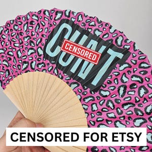 May include: A pink hand fan with a leopard print design and the word "CUNT" in large black letters with a red "CENSORED" stamp. The fan is made of wood and paper.
