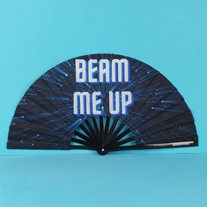 May include: A black and blue folding fan with the text "BEAM ME UP" in white and blue. The fan has a space-themed design with radiating blue lines and stars. The fan is open, displaying its full semi-circular shape.