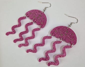 May include: A pair of pink glitter jellyfish earrings. Each earring features a scalloped, semi-circular top with small, light-colored dots and three wavy, dangling tentacles. The earrings have silver-colored fish hook closures.