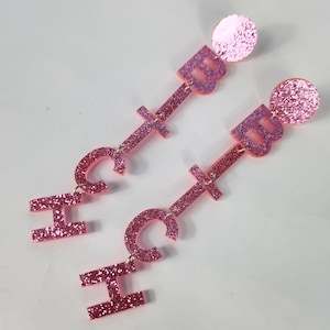 Puede incluir: Un par de pendientes colgantes de purpurina rosa con las letras "HCTB" colgando de cada pendiente. Los pendientes están hechos de acrílico y tienen un acabado de purpurina rosa.