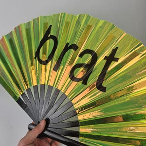 May include: A hand fan with a green and gold iridescent finish. The word "brat" is printed in large black letters across the fan. The fan's handle and ribs are black and gray.