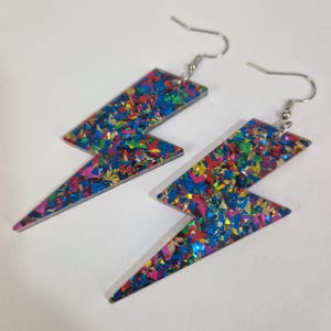 Könnte beinhalten: Ein Paar bunte Blitz-Ohrringe. Die Ohrringe sind aus einem Material mit einem Konfetti-ähnlichen Design in Blau-, Rosa-, Gelb- und Grüntönen gefertigt. Die Ohrringe haben silberfarbene Fischhaken-Ohrdrähte.