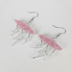 May include: A pair of dangle earrings with a pink glitter oval, clear disc, and silver glitter lightning bolt shapes. The earrings have silver hooks. The earrings are a fun, quirky accessory.