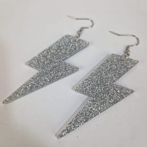 May include: A pair of silver glitter lightning bolt earrings. The earrings have a hook closure and are made of a sparkly, textured material. These earrings are a fun and fashionable accessory.