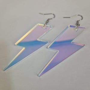 Pode incluir: Um par de brincos de raio iridescentes. Os brincos são feitos de um material transparente e reflexivo que brilha com tons de azul, roxo e amarelo. Eles têm ganchos de orelha prateados.