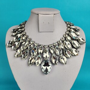 May include: A silver necklace with a large, teardrop-shaped pendant made of clear crystals. The necklace is made of many smaller crystals arranged in a cascading pattern.