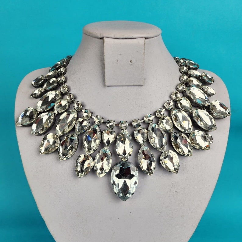 May include: A silver necklace with a large, teardrop-shaped pendant made of clear crystals. The necklace is made of many smaller crystals arranged in a cascading pattern.