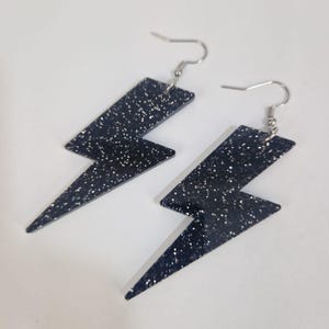 May include: A pair of dark blue glitter lightning bolt earrings with silver-colored hooks. The earrings are a bold, geometric shape, with a sparkling, speckled texture. The earrings are designed to dangle.