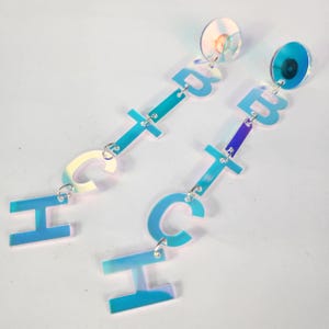 May include: A pair of iridescent blue and purple dangle earrings with the letters "H", "C", "T", and "B" hanging from each earring.