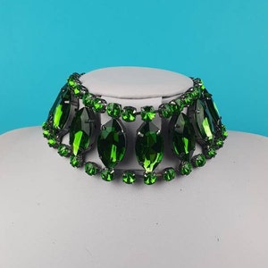 May include: A green rhinestone choker necklace with a black metal chain. The necklace is made of large, faceted rhinestones that are arranged in a cascading pattern.