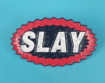 May include: A red and black enamel pin with a white glitter-filled word "SLAY" in a starburst shape.