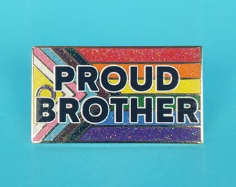 May include: A rectangular pin with a rainbow flag design and the words "PROUD BROTHER" in bold white letters. The pin has a gold border and a glittery finish, set against a light blue background.