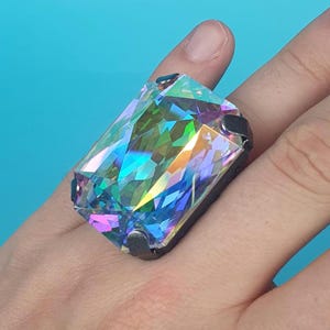 Puede incluir: Un anillo rectangular grande con una piedra preciosa facetada e iridiscente. La gema refleja un espectro de colores, incluyendo azul, verde y morado. El engaste del anillo es oscuro y rectangular, y se lleva en un dedo.