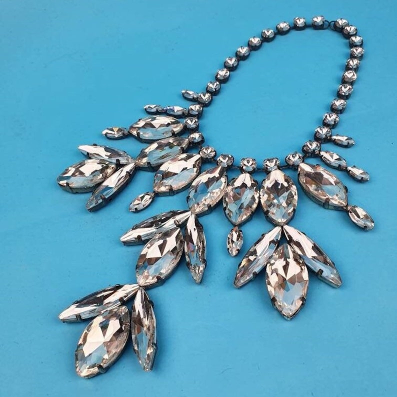 Pode incluir: Um colar de prata com um grande pingente de cristal em forma de l&aacute;grima. O pingente &eacute; cercado por cristais menores, criando um efeito de cascata. O colar est&aacute; sobre um fundo azul.