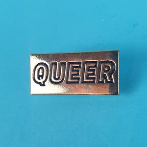 Może przedstawiać: Złota szpilka z napisem "QUEER" w czarnych, obwiedzionych literach.