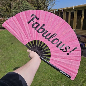 Puede incluir: Un abanico plegable de color rosa con purpurina y mango negro con la palabra "Fabulous!" impresa en negro.
