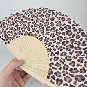 May include: A hand fan with a leopard print design. The fan features a beige wooden frame and a fabric with a pattern of brown and black spots on a cream background. The fan is open, displaying its full shape.