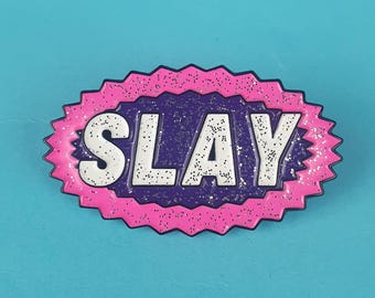 May include: A pink and purple enamel pin with a white outline and glitter. The pin says "SLAY" in all caps.