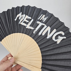 May include: A black hand fan with the words "I'M MELTING" in white lettering. The fan is made of black paper and light wood. The fan is open, displaying the pleated design.