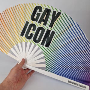 May include: A rainbow-striped hand fan with the words "GAY ICON" in large black letters. The fan has a white handle and is held open. The bottom of the fan has the text "@DRAGGEDOUTLONDON".