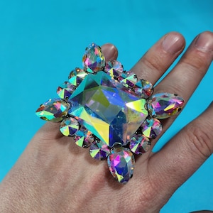 May include: A large, iridescent, square-shaped ring with a large, faceted, clear crystal in the centre. The ring is surrounded by smaller, faceted crystals.