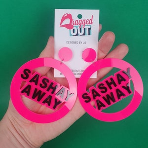 May include: A pair of pink acrylic earrings with the words "Sasha Away" in black lettering. The earrings are in the shape of a circle with a smaller circle inside.