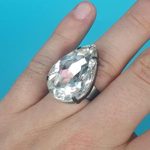 May include: A large, clear teardrop-shaped crystal ring with a silver setting. The ring is worn on a person's finger.