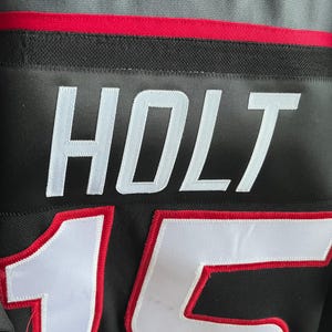 May include: Black jersey with the name "HOLT" in white block letters. Below the name is the number "15" in white with a red outline. The jersey has a red and black trim.