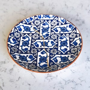 May include: A round ceramic plate with a blue and white floral pattern and a gold repair line. The plate has a brown rim and is set against a white background. The design features stylized flowers and leaves.