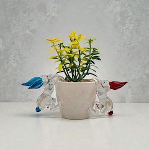 May include: A small, off-white ceramic pot with a green and yellow plant. Two clear glass rabbit figurines with blue and red ears are attached to the pot. The background is a light grey. The pot is likely measured in centimetres.