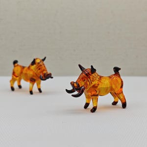 Könnte beinhalten: Zwei Wildschwein-Figuren aus bernsteinfarbenem Glas. Die Wildschweine stehen auf einer weißen Oberfläche. Die Wildschweine haben schwarze Augen, Schnauzen und Hufe.